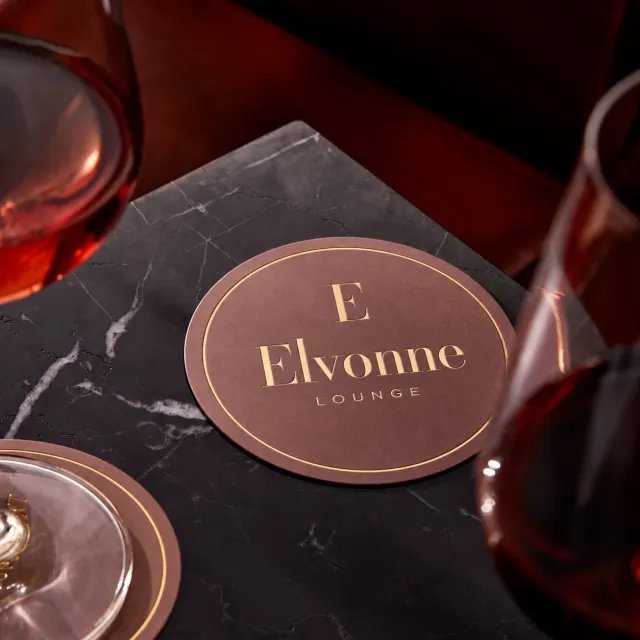 Rund ELVONNE Lounge-brikke i brunt med gullpreg på en sort marmorplate mellom to glass. KI-generert.