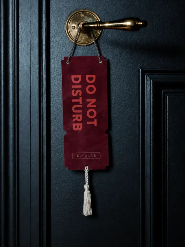 Vinrødt «Do not disturb»-skilt med ELVONNE-merke og dusk, hengt på et messinghåndtak på en mørk dør. KI-generert.