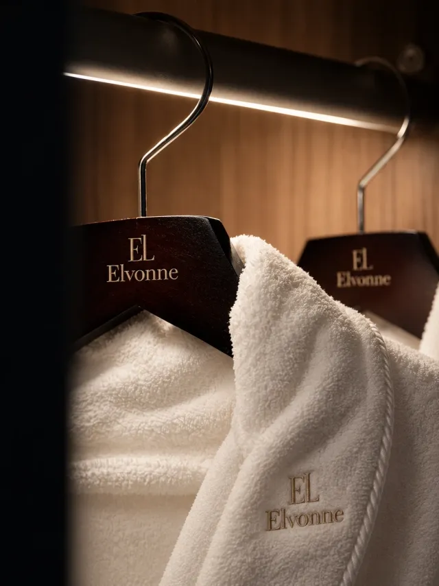 Hvite badekåper på mørke kleshengere, begge med EL-monogram og ELVONNE brodert. KI-generert.